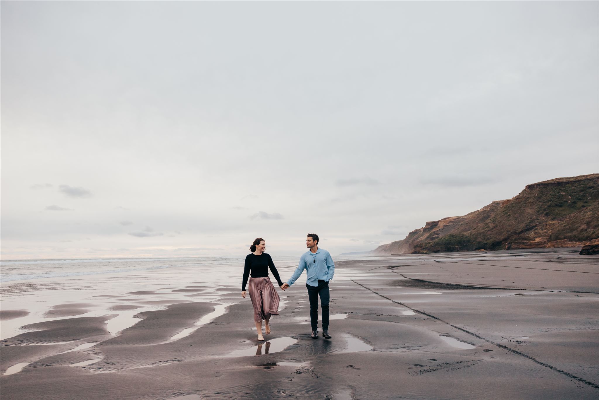 Anna & Guy walking along the beach holding hands during their engagement shoot with auckland photographer michelle kelly photoraphy