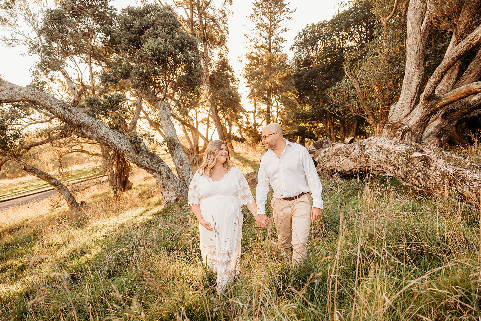 Angela and her husband walking through the beautiful long grass while the sunsets in the background and they're photographed by auckland maternity photographer michelle kelly