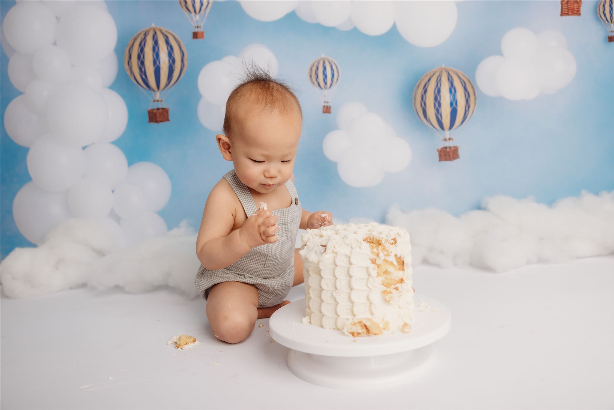 Evan eating cake wearing a cute romper with a air balloon themed backdrop during his cakesmash photoshoot to celebrate his first birthday in the studio with michelle kelly photography