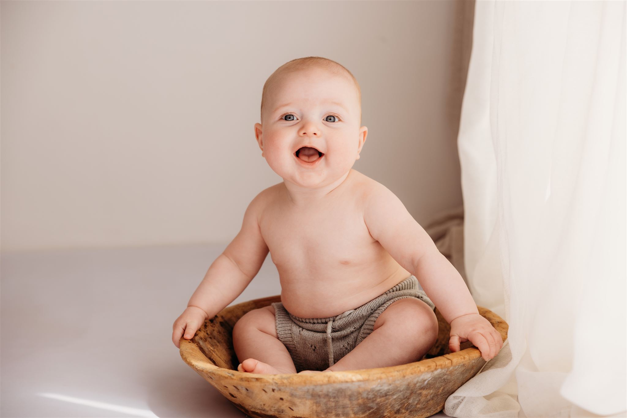 Little baby Parker sitting in a wooden bowl and giggling at the camera during his milestone photography shoot in my papakura auckland newborn studio